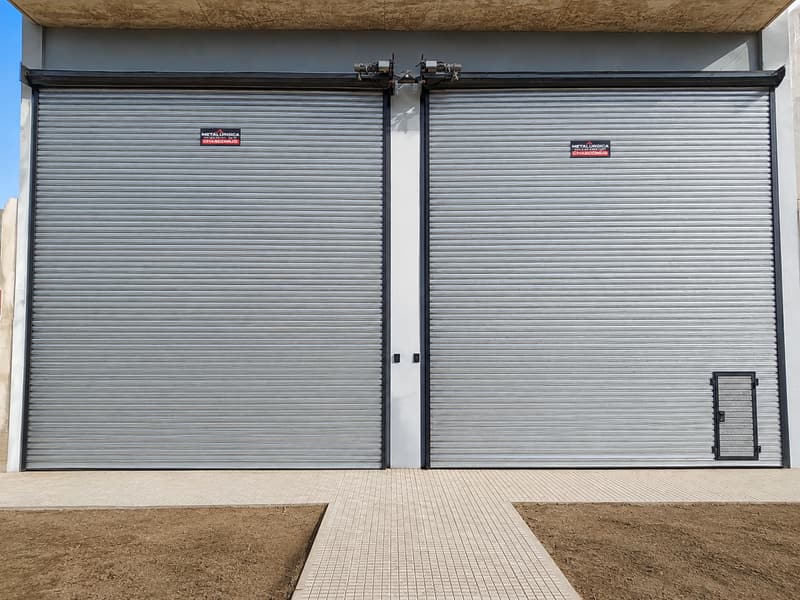 Dos cortinas metálicas de enrollar grandes con motor, una con puerta de paso. La cortina de la izquierda mide 4 m de ancho por 5,5 m de alto